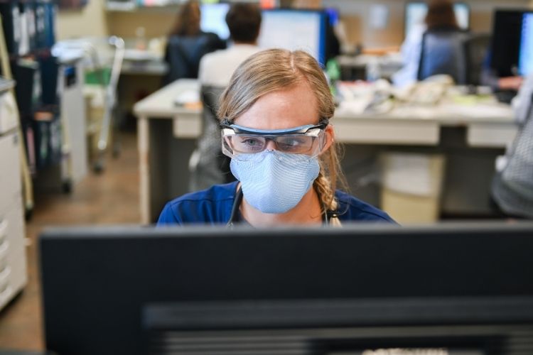 Healthcare professional wears protective gear and works at a computer.