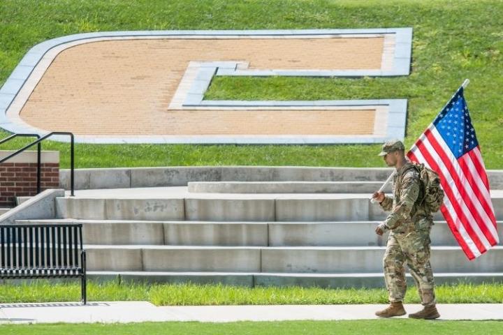A Solider holding the American Flag walking in front of the Power C on Chamberlain Field.