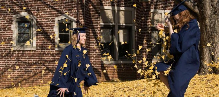 two students in graduation gowns playing in the leaves